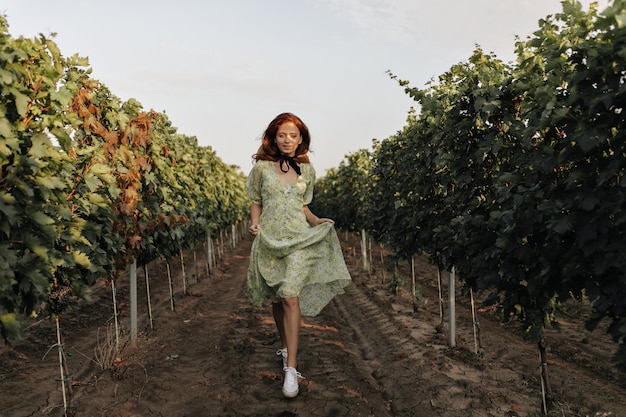 Free photo full length photo of stylish girl with foxy hairstyle and black bandage on neck in green dress walking and smiling on vineyards