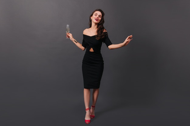 Free photo full-length photo of woman in black fitting dress and in high heels posing on black background with glass of champagne in her hands.