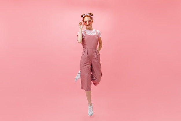 Full-length photo of woman in pink jumpsuit and accessories jumping on isolated background.