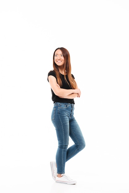 Full length photo of young pretty chinese woman in black tshirt and jeans standing with crossed hands, looking aside