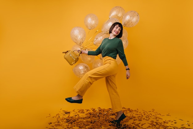 Free photo full-length portrait of adorable woman dancing with party balloons. indoor shot of blissful brunette girl in yellow pants having fun in her birthday.