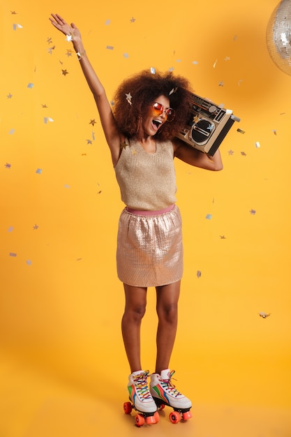 Full length portrait of beautiful overjoyed afro american disco woman with raised hand, standing on roller skates, holding boombox