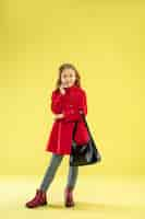 Free photo a full length portrait of a bright fashionable caucasian girl in a red raincoat holding a black bag on yellow.