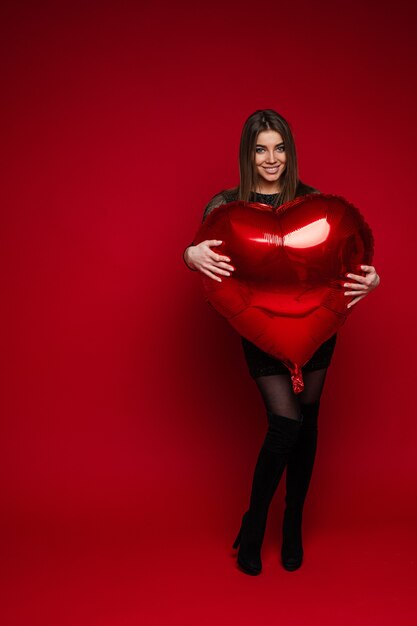 Free photo full length portrait of cheerful brunette girl in dress and boots embracing red heart-shaped balloon on red background. saint valentine concept.