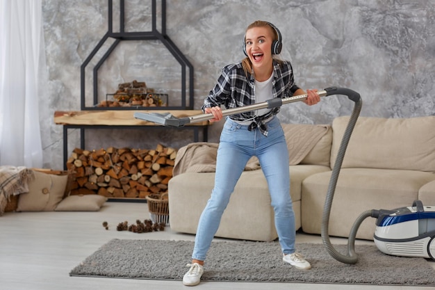 Free photo full length portrait of cheerful young woman 20s listening to music via headphones and having fun with vacuum cleaner in the house. busy, cleaning day. home, housekeeping concept