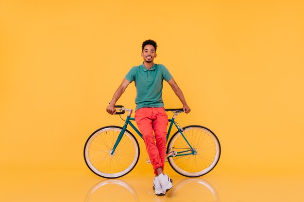 Free Photo full-length portrait of confident african man standing in front of his bike.  emotional black guy in bright outfit posing with bicycle.