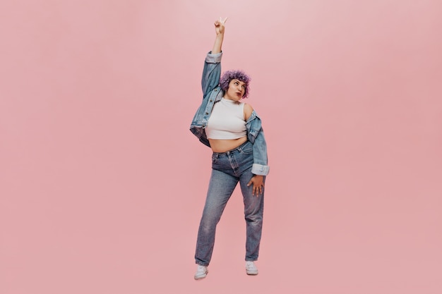 Full-length portrait of curly-haired woman with purple hair in denim fashionable suit and white sneakers.