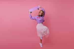 Free photo full-length portrait of debonair young lady fooling around with skateboard. indoor portrait of slim stylish girl dancing on pastel.