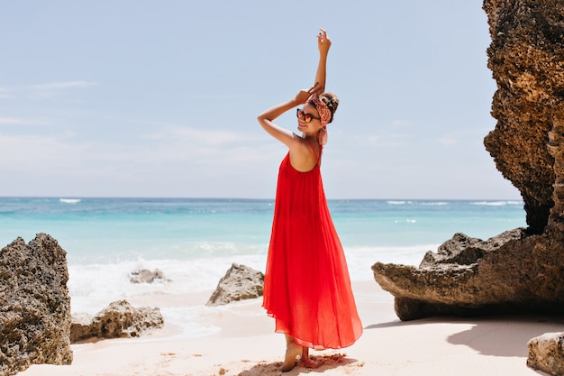 Full-length portrait of elegant european woman smiling near ocean. Stunning tanned girl in red posing in the beach