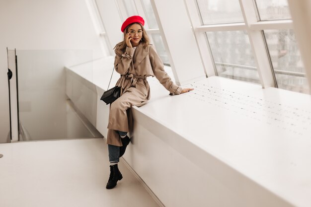 Full-length portrait of fashionable Asian woman in black shoes, jeans and light trench coat, posing by window with black bag