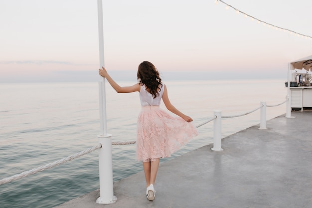 Free photo full-length portrait from back of elegant brunette girl walking along the ocean wharf and enjoying beautiful morning view