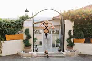 Free photo full-length portrait from back of woman in hat and striped attire standing with legs crossed in front of black iron gates