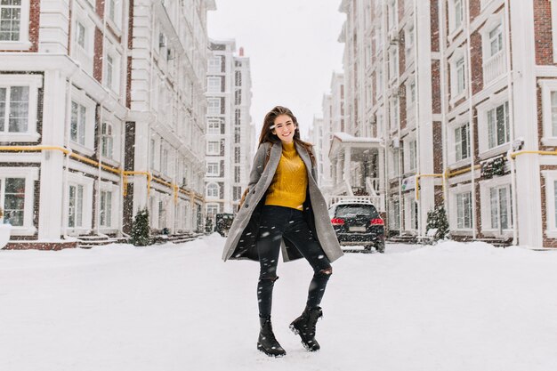 Free photo full-length portrait of graceful woman wears trendy yellow sweater fooling around on the street under snowfall. outdoor photo of slim european woman in warm coat dancing on urban street.