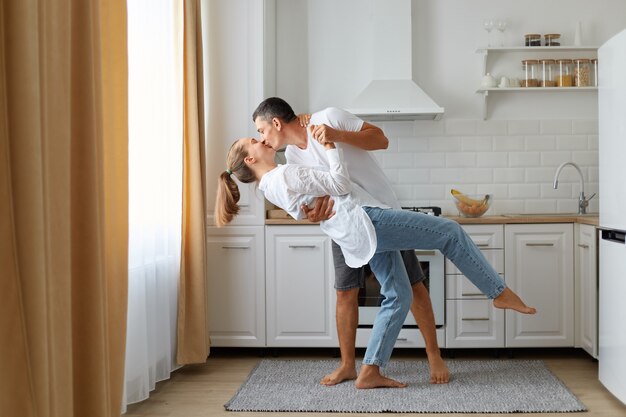 Full length portrait of happy couple wearing casual clothing dancing together in kitchen, husband kissing his wife, being happy to spend time together at home.