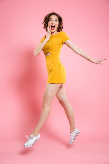 Full length portrait of happy exited woman in elegant yellow dress  while jumping over pink 