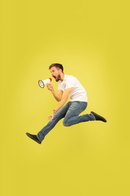 Free photo full length portrait of happy jumping man isolated on yellow background. caucasian male model in casual clothes. freedom of choices, inspiration, human emotions concept. calling with mouthpeace.