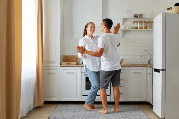 Full length portrait of happy positive falling in love couple dancing in kitchen together, spending time together at home, expressing romantic feelings.