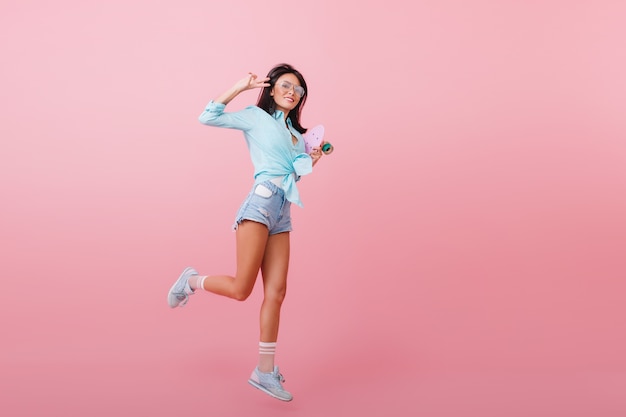 Full-length portrait of inspired dark-haired woman in blue shirt jumping and laughing. Glad hispanic girl with longboard dancing with peace sign.