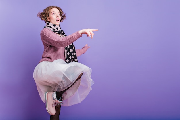 Free Photo full-length portrait of inspired girl in lush skirt posing on one leg on purple wall. graceful caucasian young woman in hat and long scarf having fun