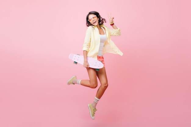 Full-length portrait of joyful dark-haired girl jumping with skateboard. Amazing hispanic lady in colorful attire dancing, holding longboard and laughing.