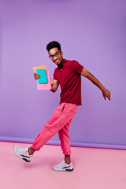 Free photo full-length portrait of joyful international student dancing after exams. smart african guy in pink pants standing with books.