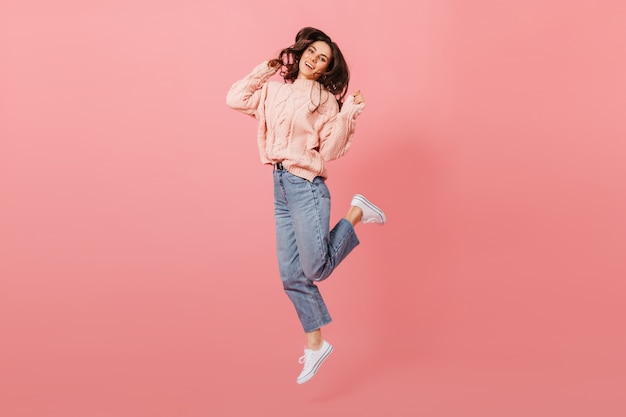 Free photo full-length portrait of jumping dark-haired girl. lady in jeans and pink sweater having fun on isolated background.