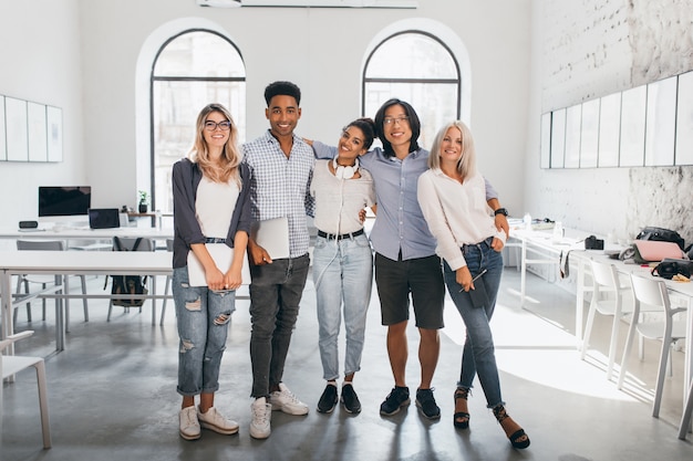 Full-length portrait of shy blonde woman in white sneakers holding laptop after seminar and stands beside african friend. Excited international students posing together after lecture in spacious hall.