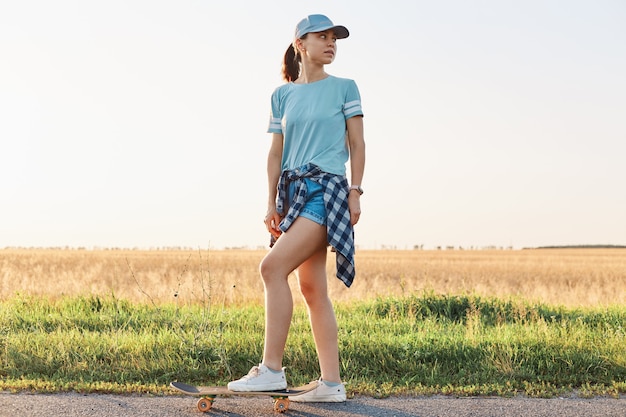 Free Photo full length portrait of slim sporty woman wearing t shirt and visor cap, standing with leg on skateboard and looking away, spending her leisure time in active way.