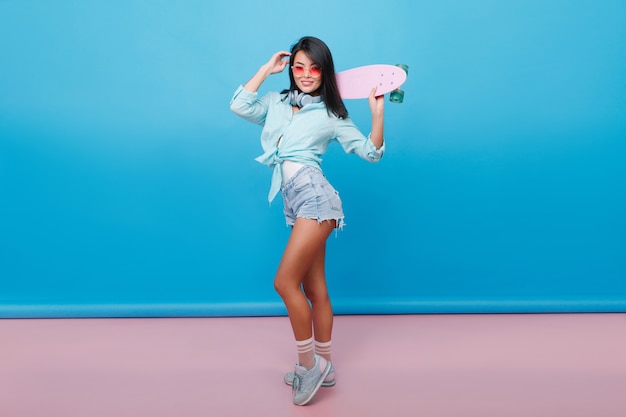 Full-length portrait of sporty girl wears cute socks and stylish cotton shirt. Slim brunette latin woman with skateboard enjoying in room with blue interior.