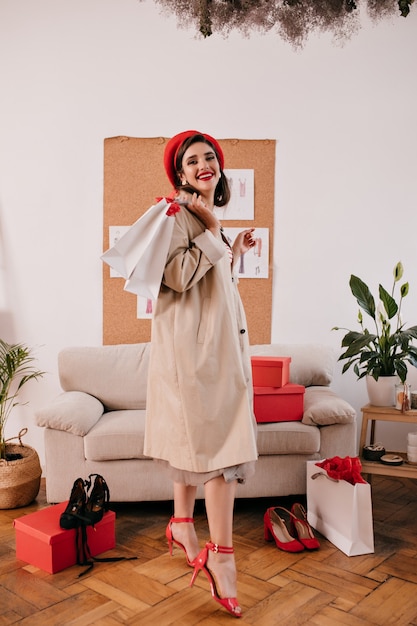 Full length portrait of woman in red beret and beige coat. Cute lady in light autumn clothes and bright hat holds purchases in apartment.