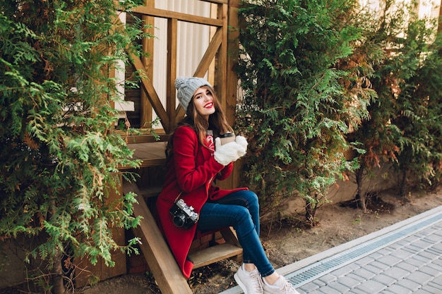 Full  length pretty girl in red coat, knitted hat and white gloves sitting on wooden stairs  between green branches outdoor. She holds coffee to go and smiling .