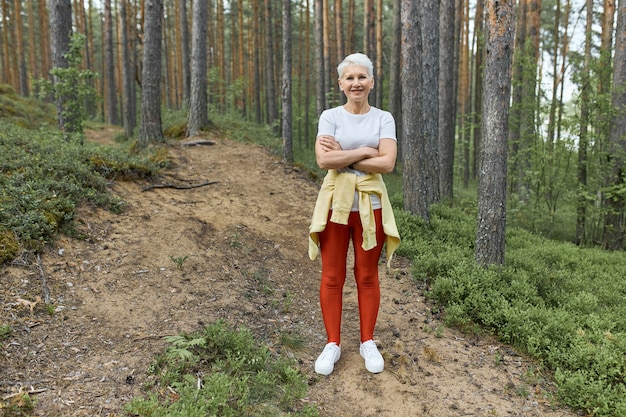 Full length shot of active mature female with blonde hair and fit body standing on trail in forest wearing sports clothes, having rest during work out, keeping arms crossed. People, activity and age