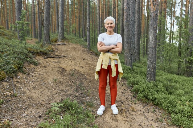 Full length shot of active mature female with blonde hair and fit body standing on trail in forest wearing sports clothes, having rest during work out, keeping arms crossed. People, activity and age