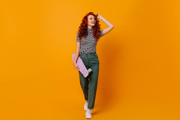 Full-length shot of curly red teenager girl with longboard in her hands. Woman in denim pants and shirt posing on orange space.