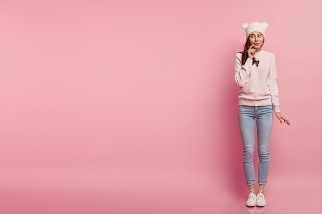 Full length shot of dreamy European woman keeps hand near lips, looks aside, has thoughtful expression, wears funny headgear with ears