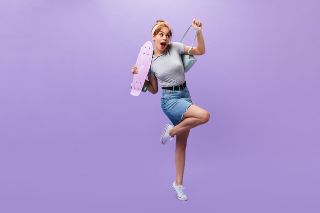 Full length shot of girl in denim skirt and shirt posing with longboard. Shy young woman in grey outfit and white modern sneakers looking into camera.