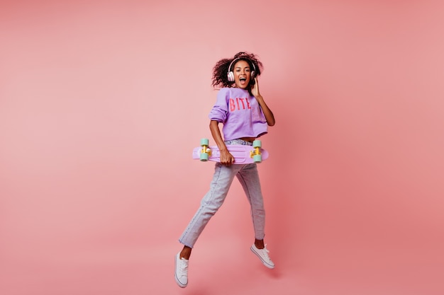 Full-length shot of stunning black woman in stylish jeans jumping on pink. curly african girl with skateboard expressing positive emotions.