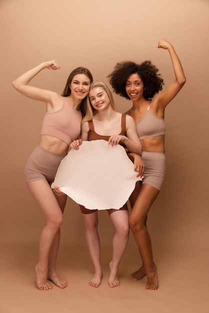 Free photo full length smiling three young interracial women holding blank placard on beige background positive emotions concept