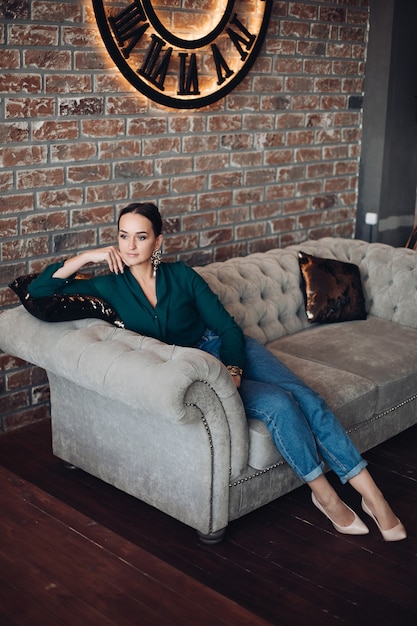 Free Photo full length stock photo of gorgeous brunette woman in casual elegant clothes and high heels relaxing on classic sofa with cushions and looking away. the room is in loft style.