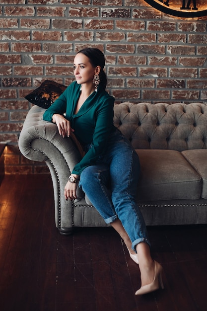 Free Photo full length stock photo of gorgeous brunette woman in casual elegant clothes and high heels relaxing on classic sofa with cushions and looking away. the room is in loft style.