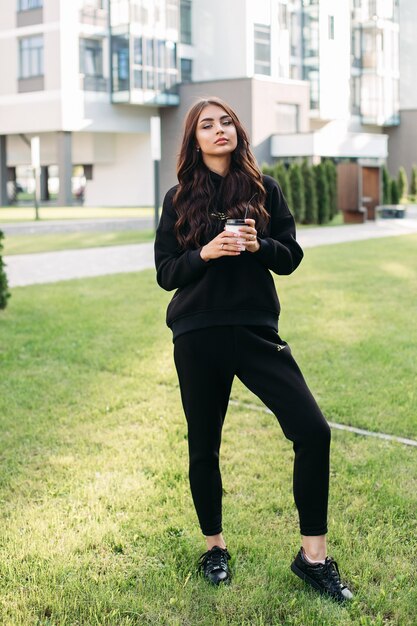 Full length stock photo of a trendy young girl with coffee wearing black sweatshirt and joggers and black sneakers on green lawn in the city.