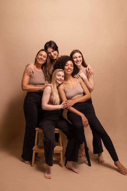 Full length stylish diverse women of caucasian and african appearance smiling at camera on beige background