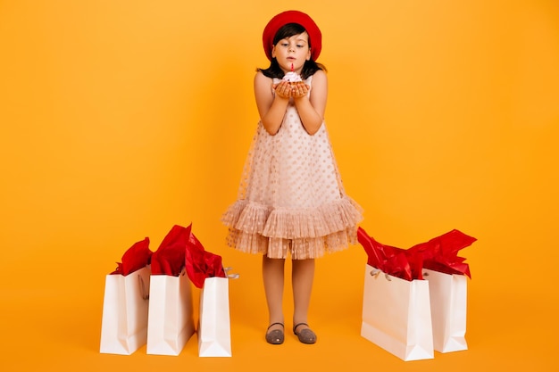 Full length view of child in dress celebrating birthday Studio shot of kid in beret blows out candle on cake