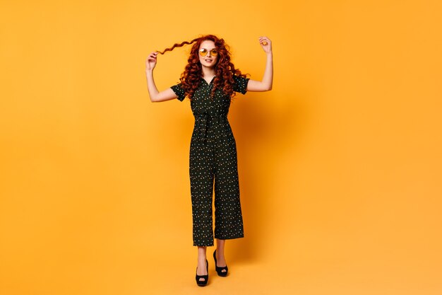 Full length view of elegant girl playing with ginger hair. Studio shot of joyful caucasian woman standing on yellow background.