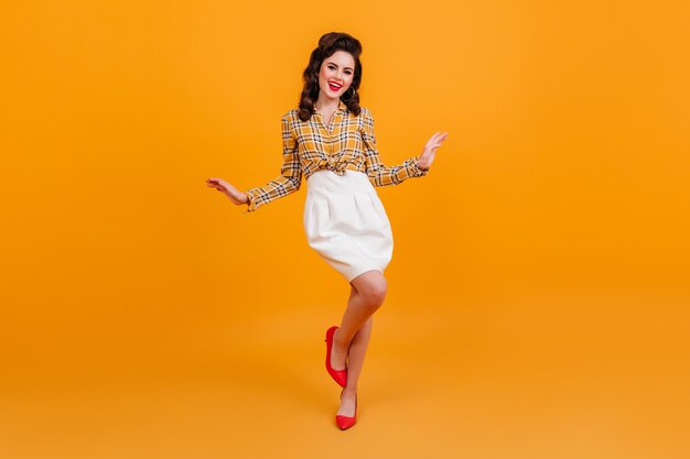 Full length view of graceful curly woman in white skirt. Pinup girl in checkered shirt dancing on yellow background.