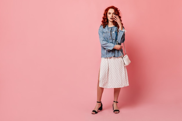 Free photo full length view of shocked ginger woman in high-heeled shoes. studio shot of amazed girl in denim jacket standing on pink background.