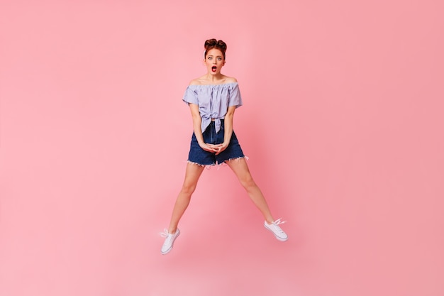 Free Photo full length view of shocked woman in denim shorts and blouse. studio shot of amazed pinup girl jumping on pink space.