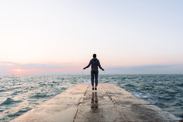 Free photo full length view of young man in sportswear jumping with skipping rope, workout on pier