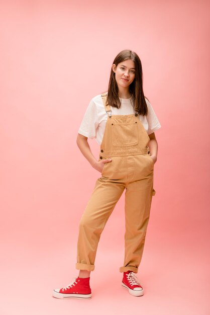 Full length young caucasian brunette woman wears tshirt overalls and running shoes on pink background Lifestyle concept