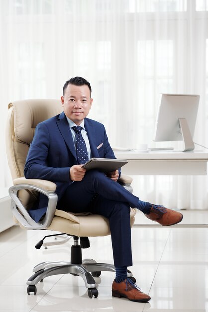 Full shot of successful Asian business executive sitting cross-legged on his luxury boss chair in light spacious office